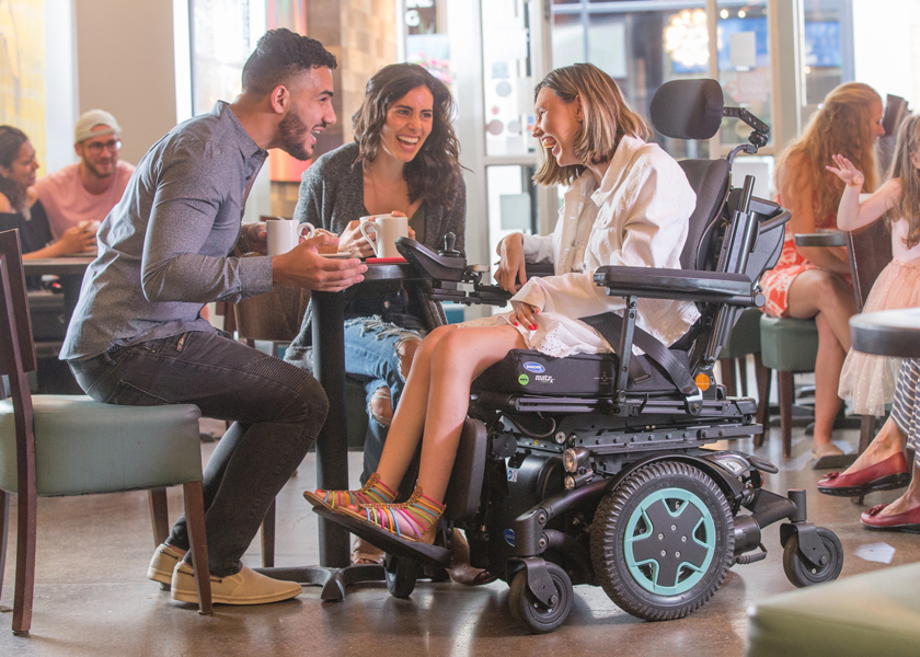 A woman using a TDX SP2 power chair laughs in a cafe with her friends A woman using a TDX SP2 power chair laughs in a cafe with her friends