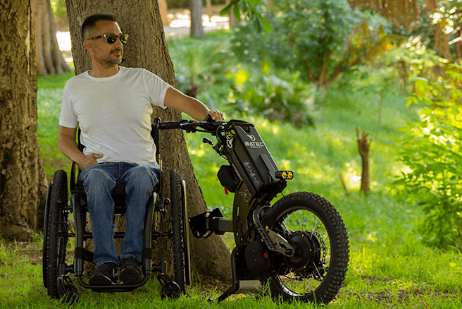 A man using a manual wheelchair sits in a verdant park. His Batec Scrambler electric handbike is detached, positioned beside him.
