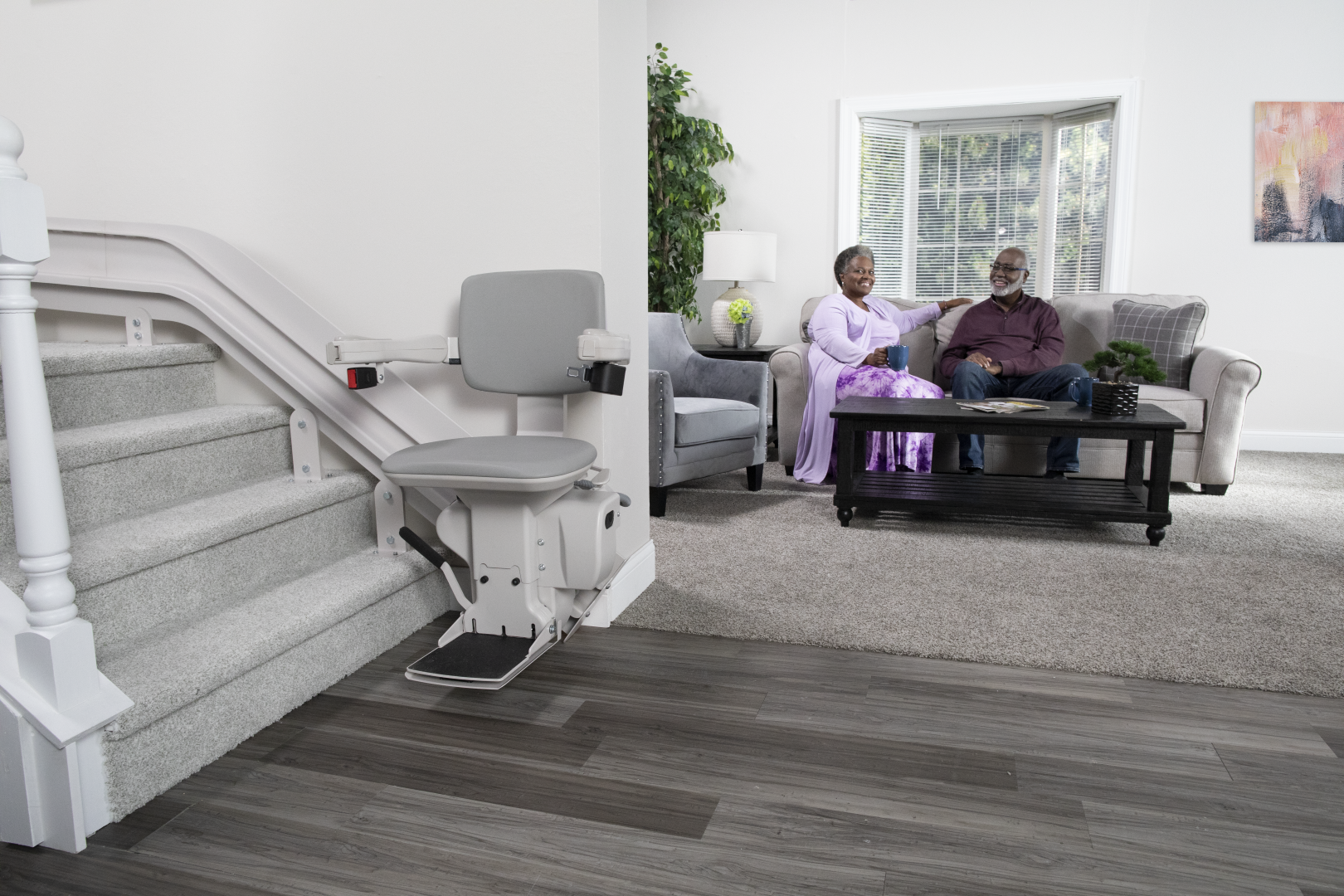 Bruno Elite curved stairlift lifestyle shot The Bruno Elite curved stairlift with the chair at the bottom of the stairs. A senior couple is relaxing in the living room nearby.