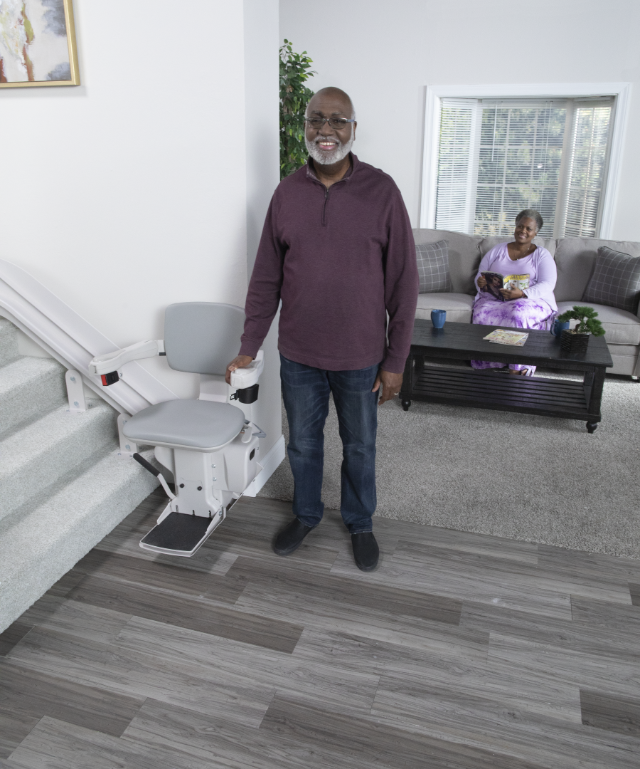 Bruno Elite curved stairlift with man beside it The Bruno Elite curved stairlift with a man standing beside it. His wife is relaxing in the living room behind him.