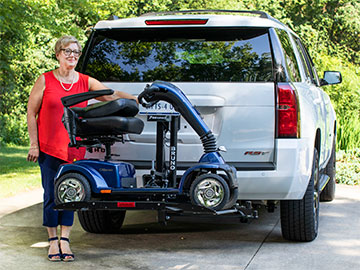 An Out-Sider scooter lift is lifted on the back of an SUV with a woman standing beside it, smiling.
