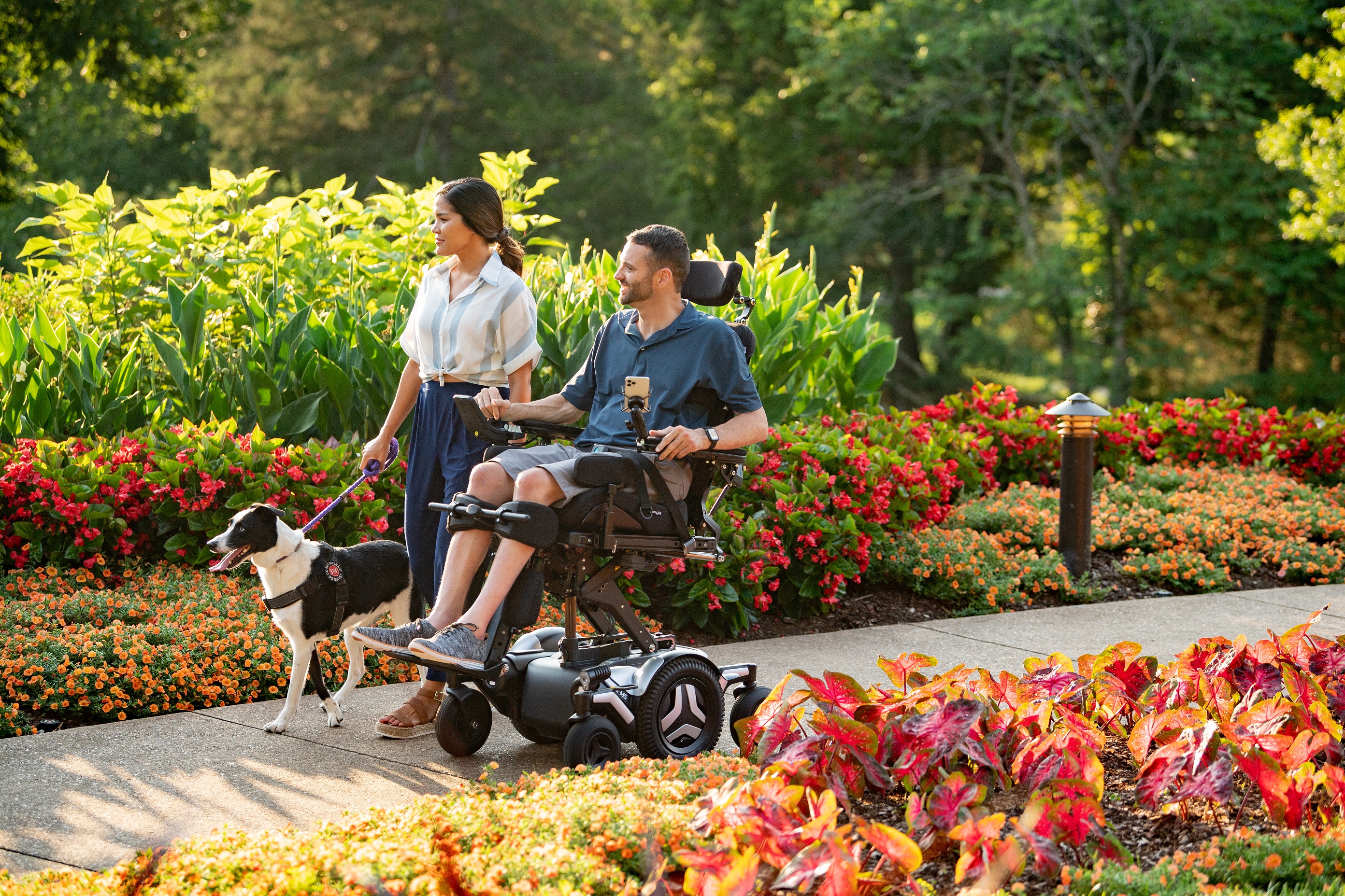 Corpus M5 couple walk dog in a flowery park A Caucasian man in a Corpus M5 chair rolls beside an Asian woman who is holding the harness of a black and white service dog. They are traveling down a path in a park filled with flowers.