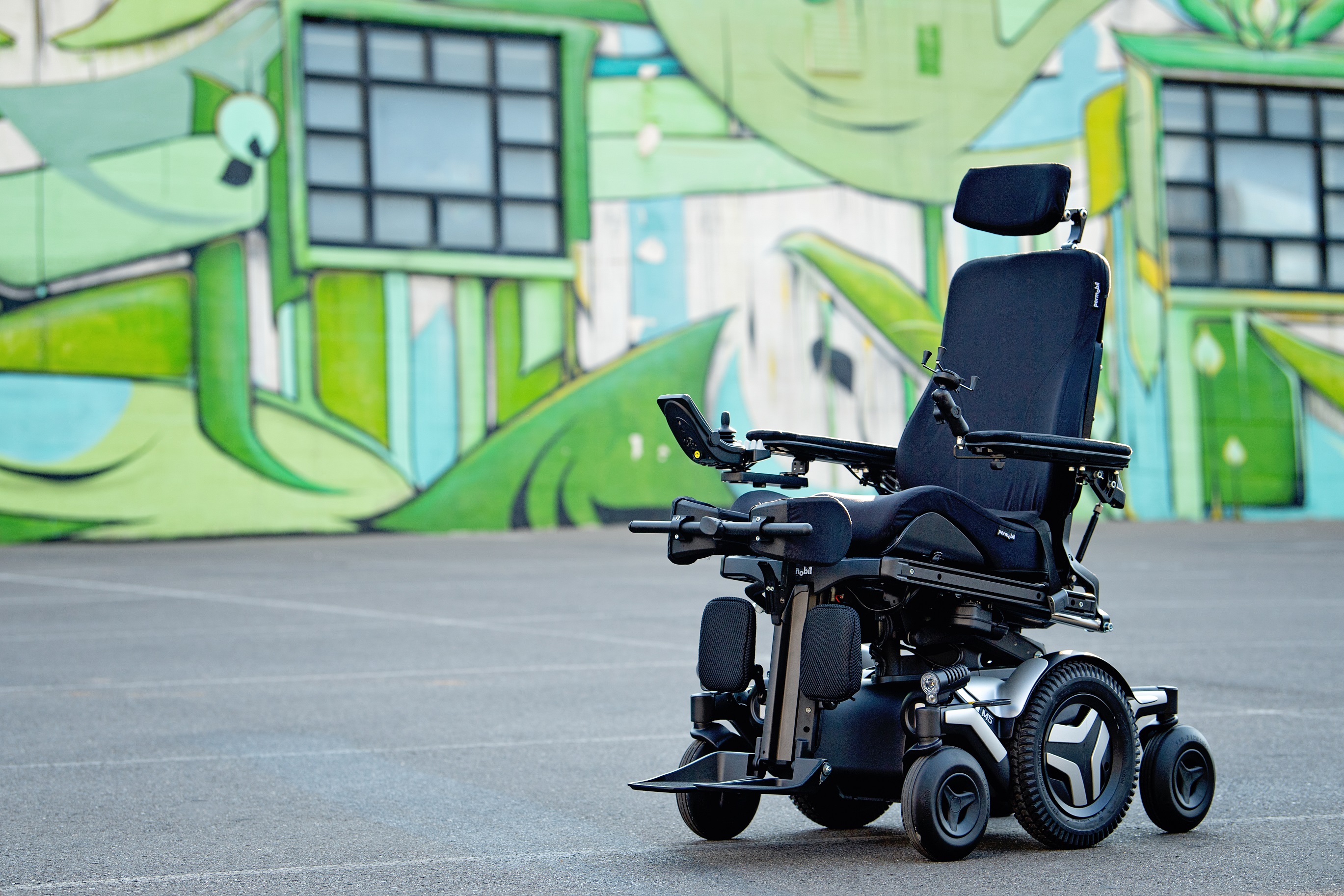 The Corpus M5 power chair with white accents, rehab seating and multiple user controls. It sits in front of a warehouse painted with urban artwork.
