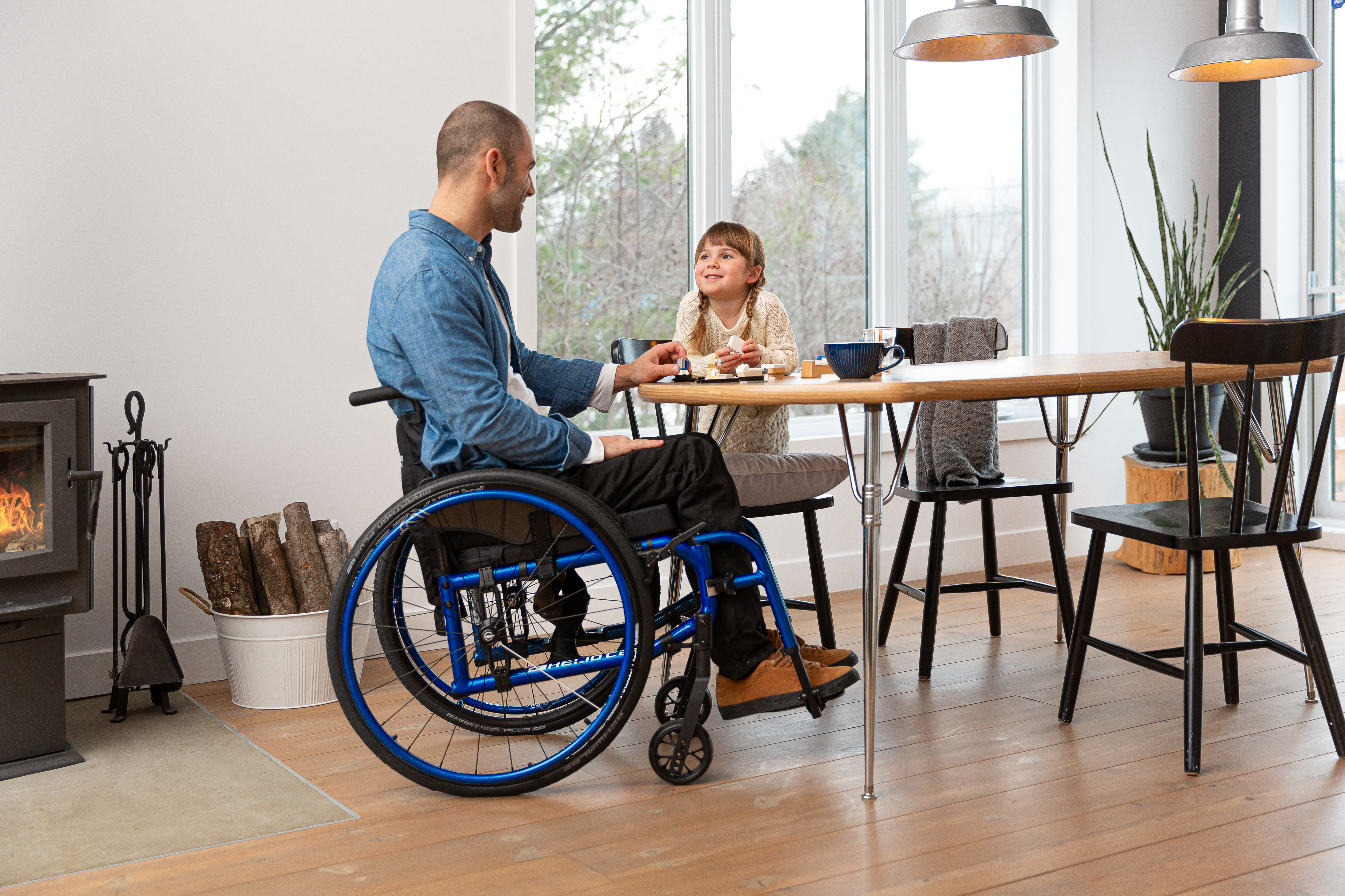 A man using a blue Helio C2 lightweight wheelchair sits at a kitchen table, smiling at his daughter. She smiles back while kneeling on a kitchen chair. There is a fireplace in the corner with a warm fire burning, and a window to the trees behind her.