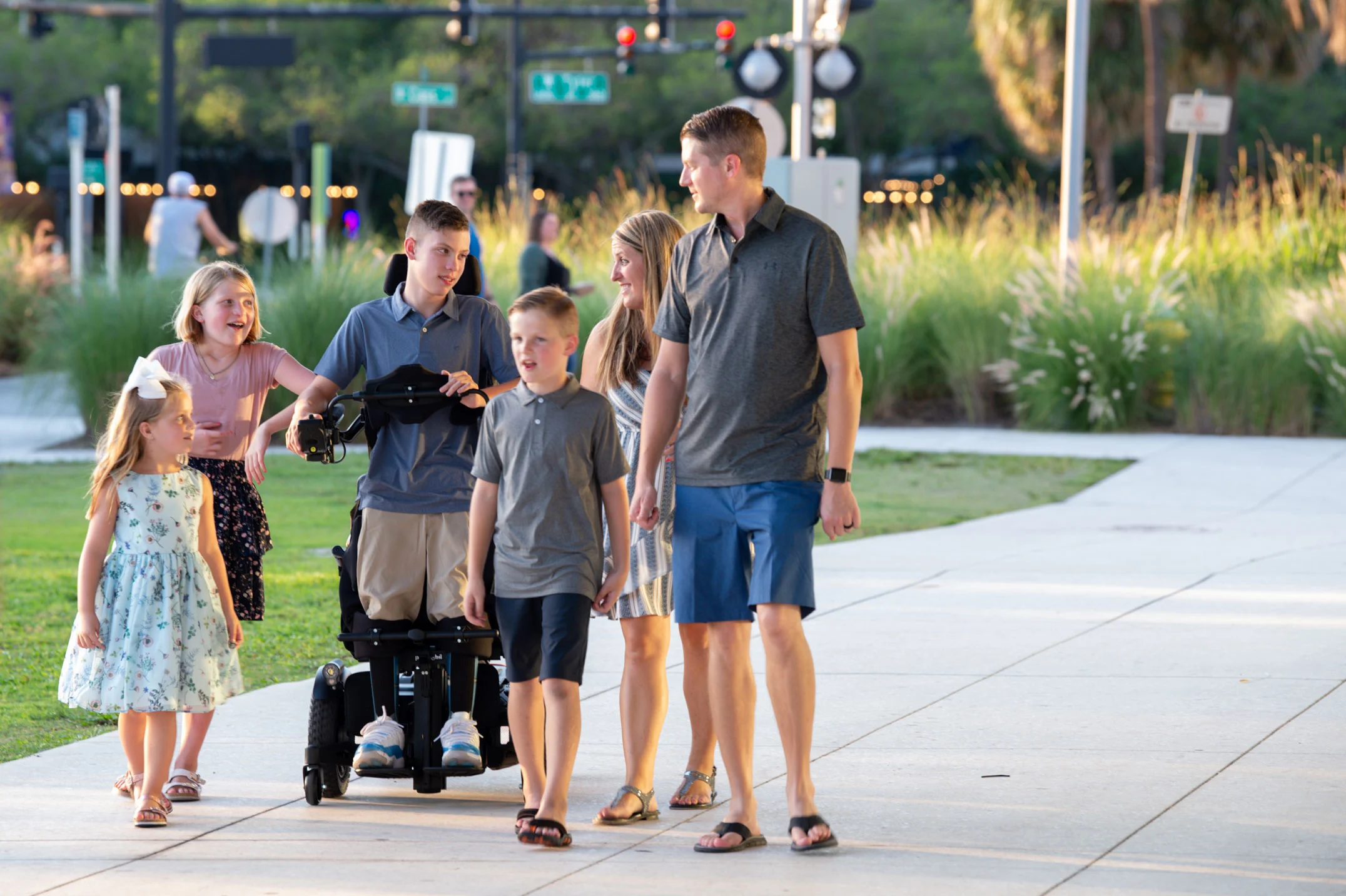 Landon, a young Caucasian man, uses his Permobil F5 VS standing wheelchair while rolling with his family in a park. They are on a cement pathway with tall grass and a street scene in the background. His family are looking at him and smiling.