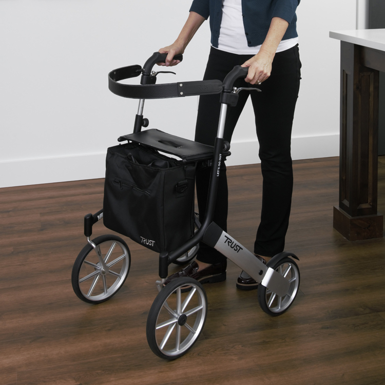 The Let's Go Out rollator sits on a dark wood laminate floor in a kitchen. A woman's hands are holding onto the handles. She is wearing shiny shoes.