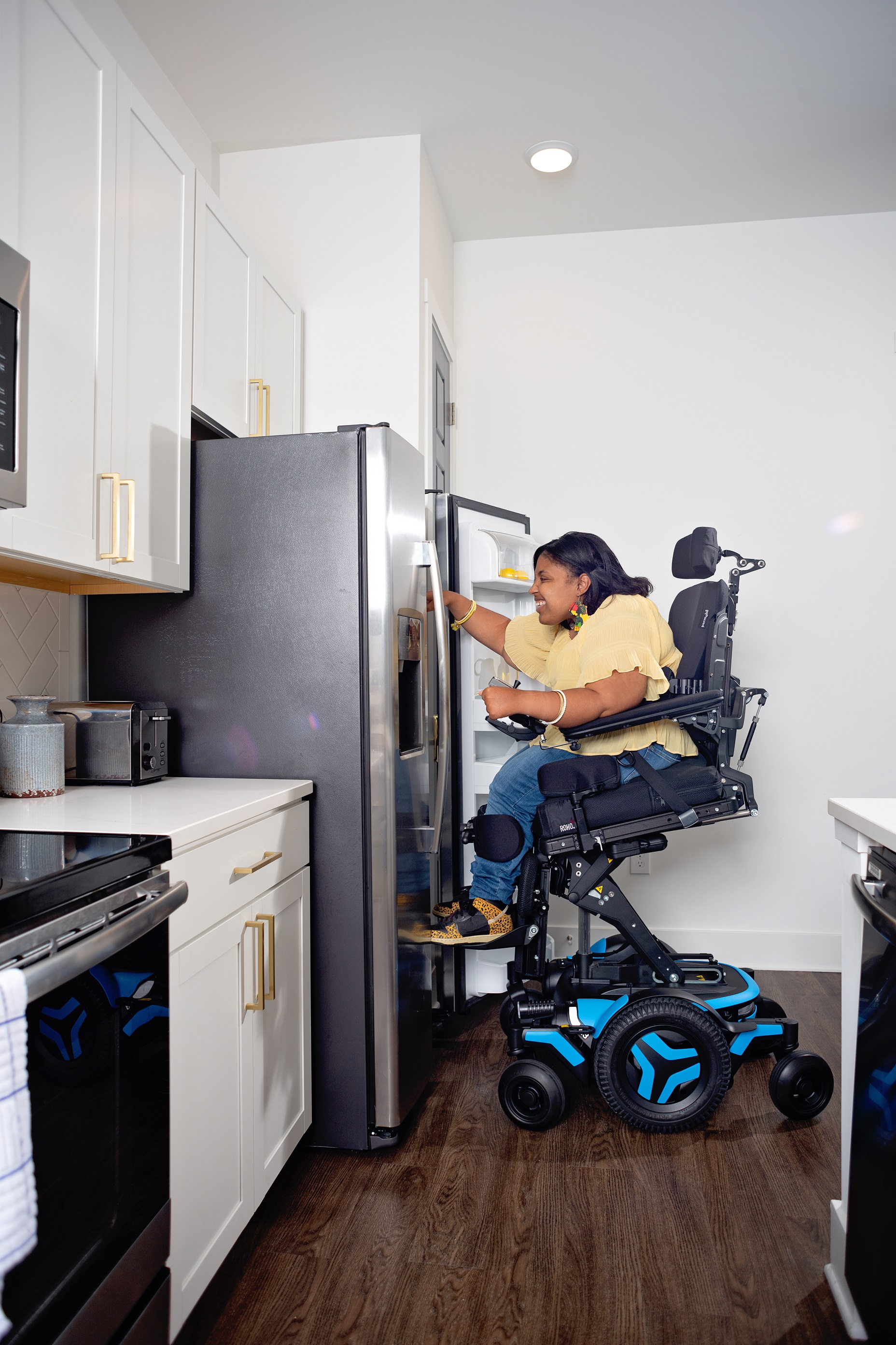 A Black woman in an M3 power wheelchair is elevated and tilted forward 20 degrees, using the ActiveReach feature to access the top shelf of her stainless steel fridge.
