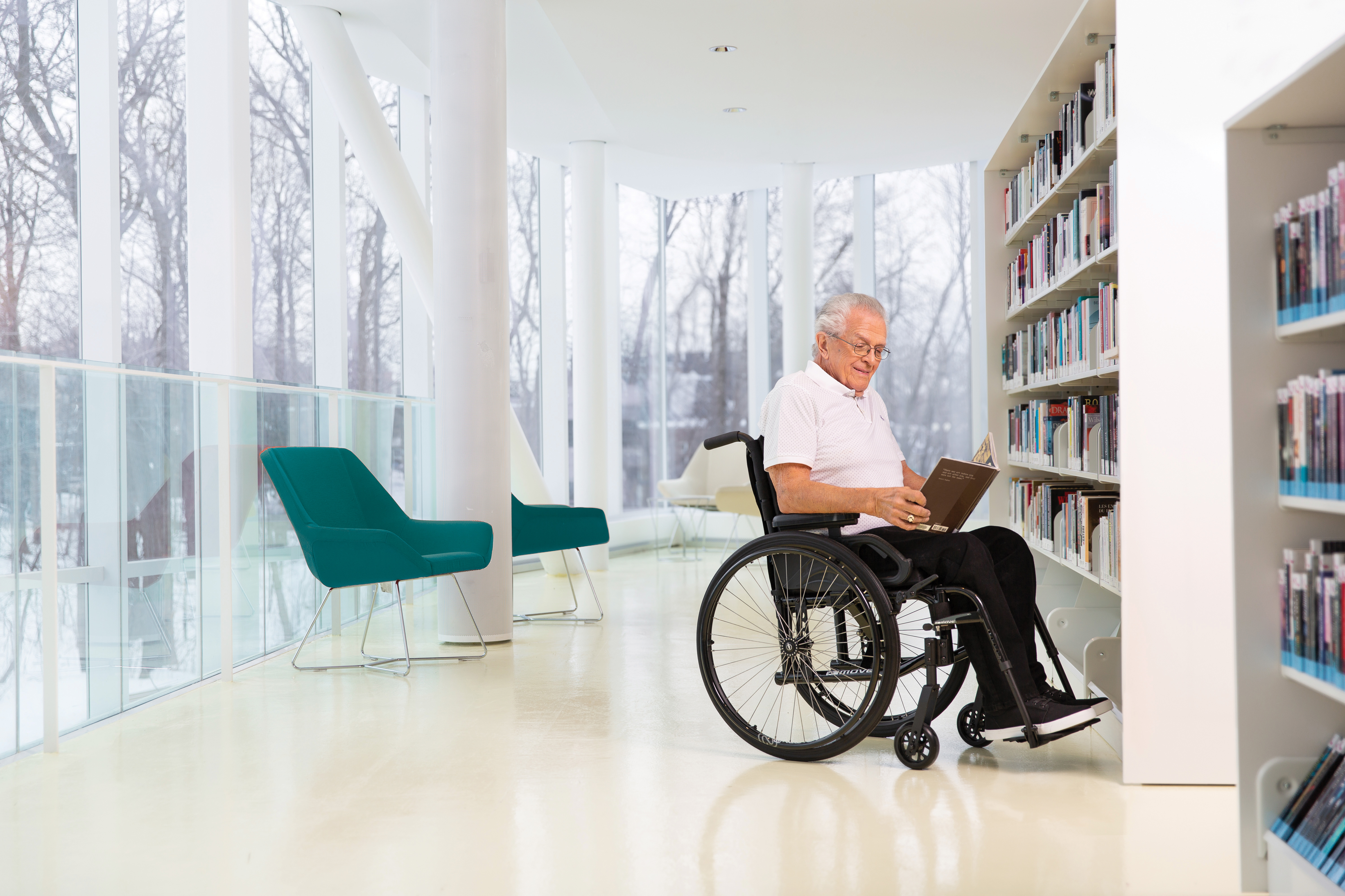 A senior man sits smiling in his Move wheelchair. He's reading a book in a brightly lit space in front of a shelf of books. There are windows behind him with a view to leafless trees. The space is painted white with pillars visible in the distance.