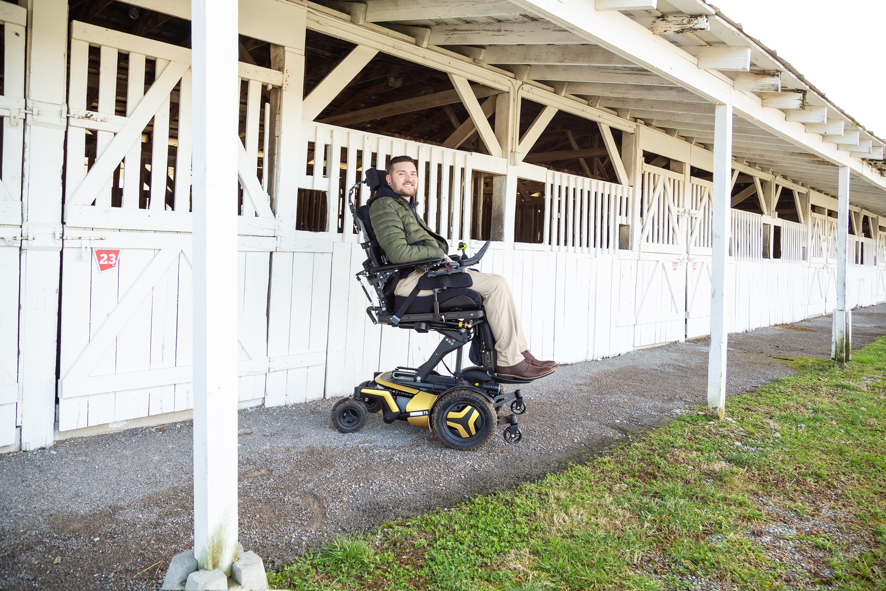 Permobil F5 Corpus standing wheelchair user outside horse stables A young Caucasian man with a bears sits in his F5 Corpus VS standing wheelchair in the elevated position. The wheelchair has yellow accents and black rehab seating. The man is in front of a row of rustic wooden horse stables, painted white.