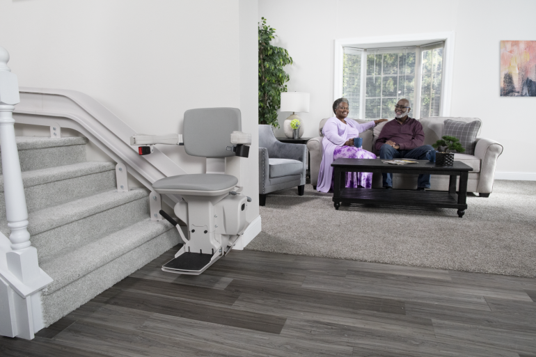 The Bruno Elite curved stairlift with the chair at the bottom of the stairs. A senior couple is relaxing in the living room nearby.