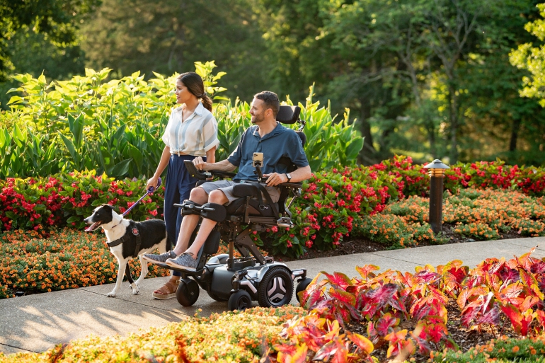 A Caucasian man in a Corpus M5 chair rolls beside an Asian woman who is holding the harness of a black and white service dog. They are traveling down a path in a park filled with flowers.