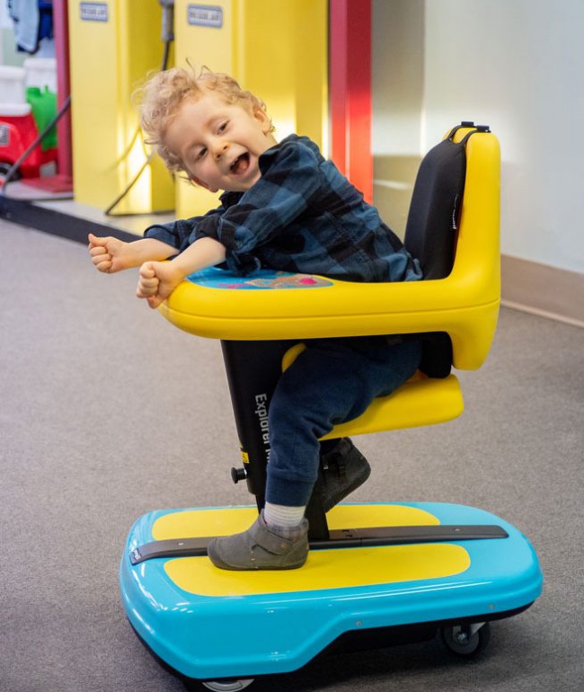 A young child, David, sits in their Explorer Mini, leaning forward and smiling. They are wearing a green and black plaid shirt, grey booties and have blonde curly hair.