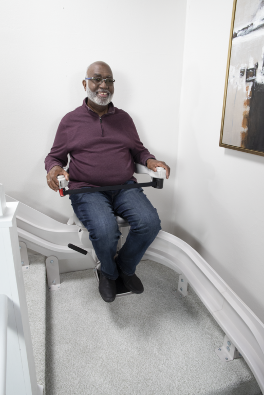 A man sits on the chair of his stairlift as he rides down a white staircase. He is smiling.