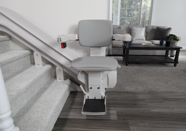 The chair of the Elite indoor curved stairlift at the bottom of a staircase, with a tidy living room in the background