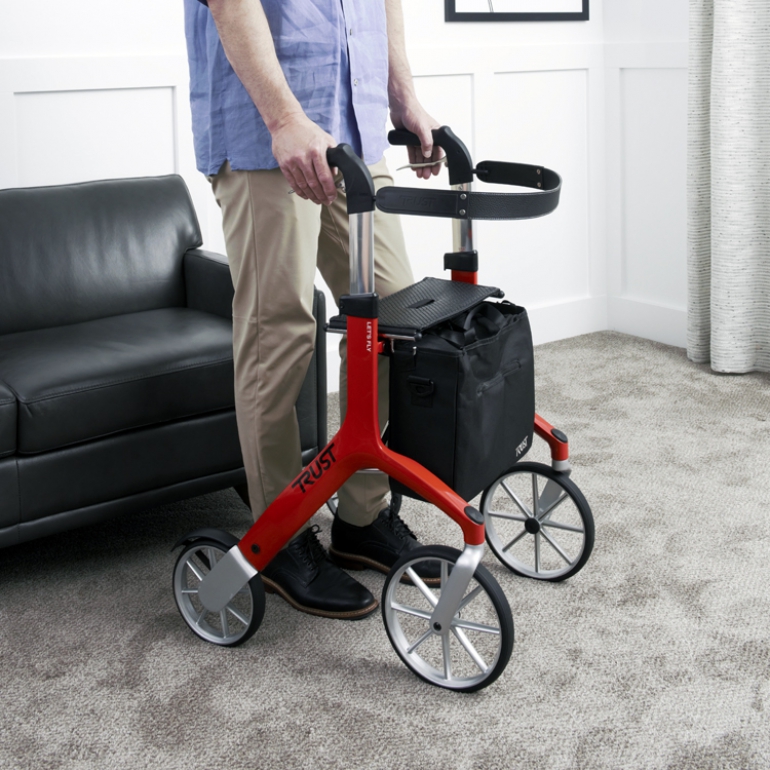 The Let's Fly Outdoor Rollator is in a carpeted living room. It is red and a man's lower torso & legs are standing behind it. His hands are gripping the handles.
