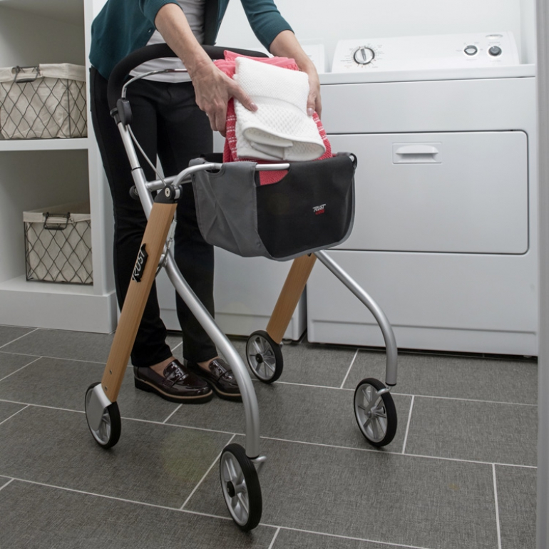 The Let's Go Indoor Rollator in the Beech colour is in a laundry room. A person's lower torso and legs are in the image, and the person is placing folded laundry into the walker's basket. A clothes dryer is visible in the background.