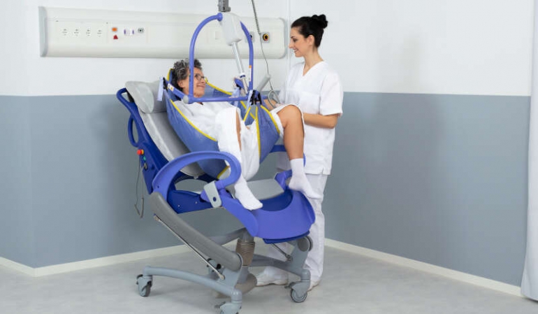 A woman is being lifted with assistance from a nurse using a Maxi Sky 2 ceiling lift.