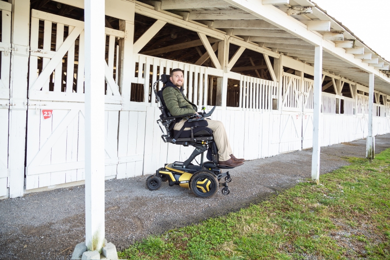 A young Caucasian man with a bears sits in his F5 Corpus VS standing wheelchair in the elevated position. The wheelchair has yellow accents and black rehab seating. The man is in front of a row of rustic wooden horse stables, painted white.