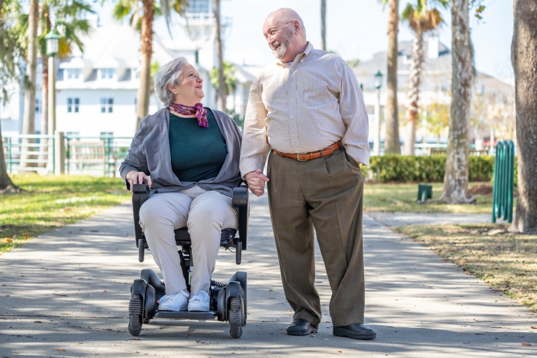 A senior couple strolls hand in hand down a path dappled with sunlight. The woman uses a WHILL power chair. They are smiling at each other.