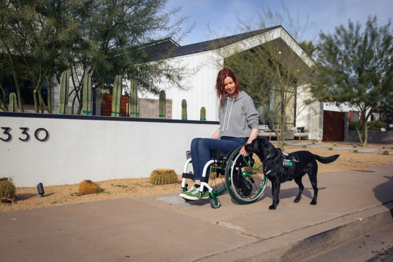 A young woman using a manual wheelchair with a SmartDrive rolls down a sidewalk in a desert town. Her black lab service dog walk at her side as she gazes down at him.