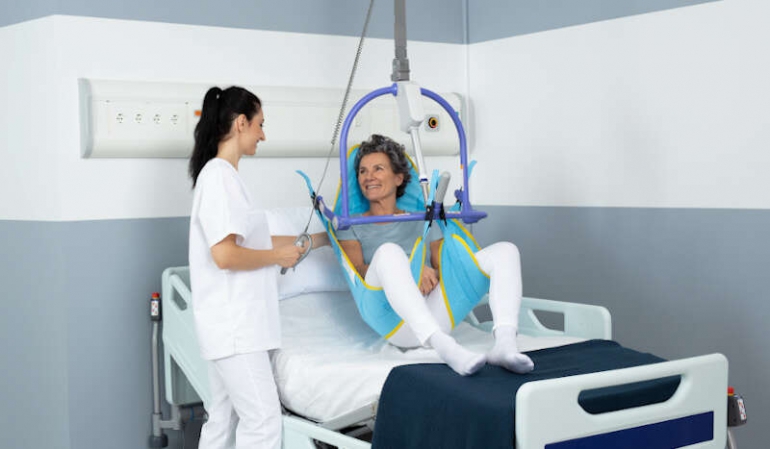 A smiling woman is being lifted from her hospital bed with a caregiver's assistance, using the Maxi Sky 2 ceiling lift