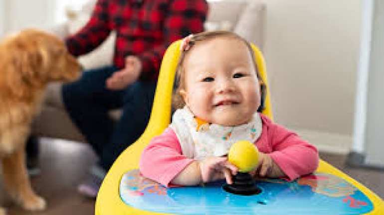 A baby sits in their Explorer Mini, facing the camera and smiling. They are wearing a pink shirt with a white bib on top. A golden retriever and a person in a red plaid shirt are visible in the background.