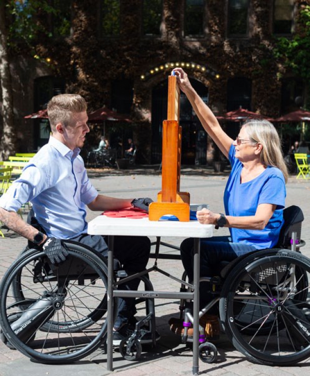 Two people using SmartDrives playing a board game at an outdoor table in the sunshine.