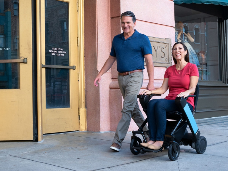 A Caucasian man and woman are out for a stroll on a city street. The woman uses a WHILL folding power chair. They are very happy.