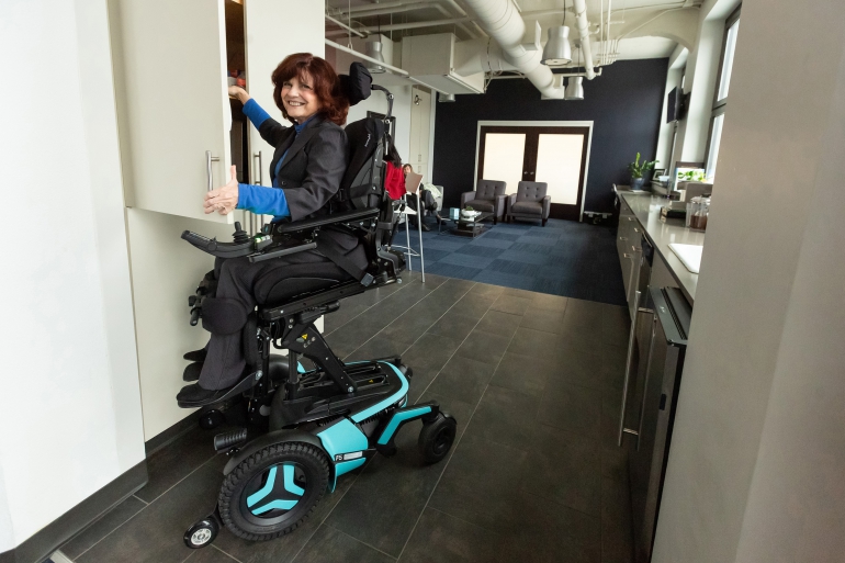 A Caucasian woman wearing a dark grey suit and a royal blue mock neck shirt uses her Corpus F5 power chair. It is in the ActiveReach position and she is reaching into a high cupboard while smiling at the camera. A living room area is behind her.