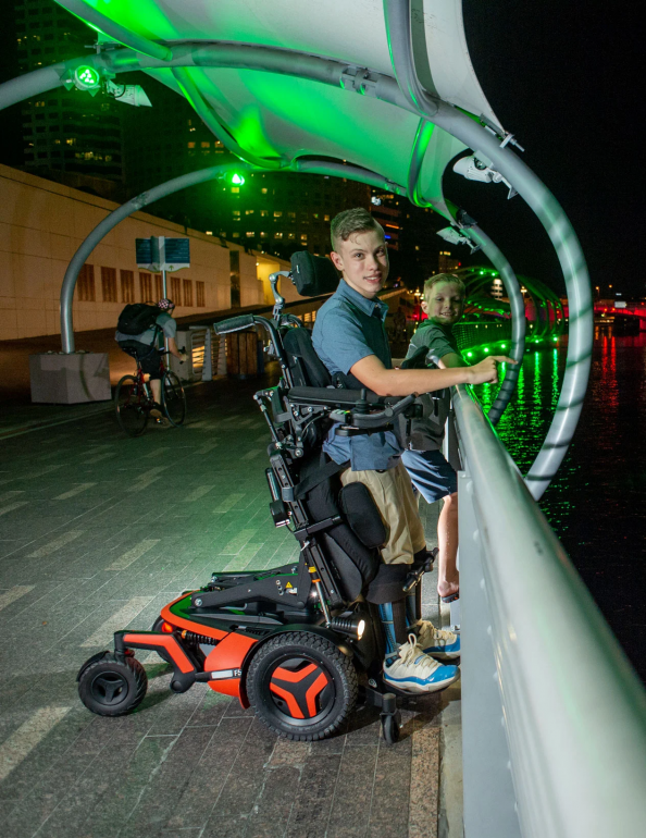 A young man using an F5 Corpus VS standing wheelchair holds his smaller brother up to the railings of a bridge to view the river below. It's nighttime and they are both smiling towards the camera.