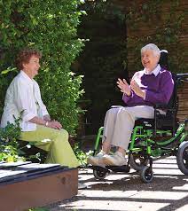 Two senior ladies sit out in the sunshine, smiling and talking. One uses a wheelchair with a Matrx Multi cushion.