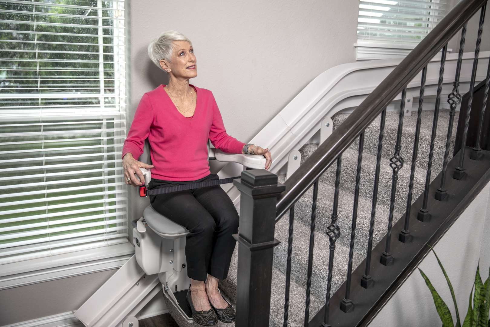 The Bruno Elite curved stairlift with a senior woman riding it down the stairs. A senior woman in a bright pink shirt rides the Bruno Elite stairlift down her stairs.