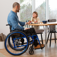 A man using a blue Helio C2 lightweight wheelchair sits at a kitchen table, smiling at his daughter. She smiles back while kneeling on a kitchen chair. There is a fireplace in the corner with a warm fire burning, and a window to the trees behind her. thumbnail