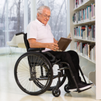 A senior man sits smiling in his Move wheelchair. He's reading a book in a brightly lit space in front of a shelf of books. There are windows behind him with a view to leafless trees. The space is painted white with pillars visible in the distance.