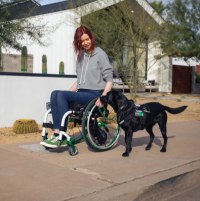 A young woman using a manual wheelchair with a SmartDrive rolls down a sidewalk in a desert town. Her black lab service dog walk at her side as she gazes down at him. thumbnail