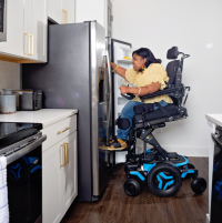 A Black woman in an M3 power wheelchair is elevated and tilted forward 20 degrees, using the ActiveReach feature to access the top shelf of her stainless steel fridge. thumbnail