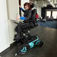A Caucasian woman wearing a dark grey suit and a royal blue mock neck shirt uses her Corpus F5 power chair. It is in the ActiveReach position and she is reaching into a high cupboard while smiling at the camera. A living room area is behind her.