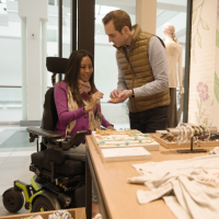 An Asian woman using a Corpus F3 power chair shops for jewelry with a Caucasian man. She is wearing a pink sweater and jeans. She smiles while the man holds some jewelry for her to look at. The man wears a gold puffer vest and black jeans. thumbnail