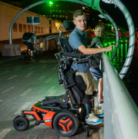 A young man using an F5 Corpus VS standing wheelchair holds his smaller brother up to the railings of a bridge to view the river below. It's nighttime and they are both smiling towards the camera.