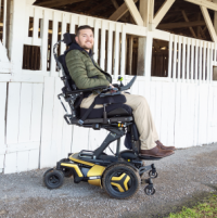 A young Caucasian man with a bears sits in his F5 Corpus VS standing wheelchair in the elevated position. The wheelchair has yellow accents and black rehab seating. The man is in front of a row of rustic wooden horse stables, painted white. thumbnail