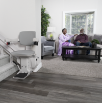 The Bruno Elite curved stairlift with the chair at the bottom of the stairs. A senior couple is relaxing in the living room nearby. thumbnail