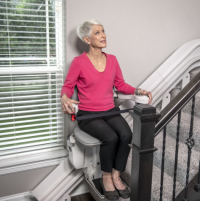 A senior woman in a bright pink shirt rides the Bruno Elite stairlift down her stairs. thumbnail