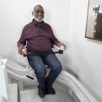 A man sits on the chair of his stairlift as he rides down a white staircase. He is smiling.