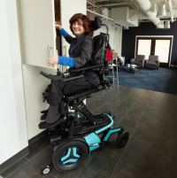 A Caucasian woman smiles and faces the camera. She is using her F5 power chair with light blue accents in ActiveReach position to access her upper cabinets. thumbnail