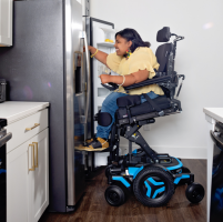 A Black woman in an M3 power wheelchair is elevated and tilted forward 20 degrees, using the ActiveReach feature to access the top shelf of her stainless steel fridge. thumbnail