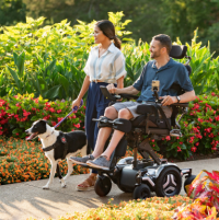 A Caucasian man in a Corpus M5 chair rolls beside an Asian woman who is holding the harness of a black and white service dog. They are traveling down a path in a park filled with flowers. thumbnail