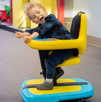 A young child, David, sits in their Explorer Mini, leaning forward and smiling. They are wearing a green and black plaid shirt, grey booties and have blonde curly hair. thumbnail
