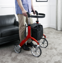 The Let's Fly Outdoor Rollator is in a carpeted living room. It is red and a man's lower torso & legs are standing behind it. His hands are gripping the handles. thumbnail