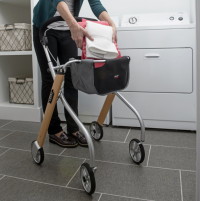 The Let's Go Indoor Rollator in the Beech colour is in a laundry room. A person's lower torso and legs are in the image, and the person is placing folded laundry into the walker's basket. A clothes dryer is visible in the background. thumbnail