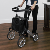 The Let's Go Out rollator sits on a dark wood laminate floor in a kitchen. A woman's hands are holding onto the handles. She is wearing shiny shoes. thumbnail